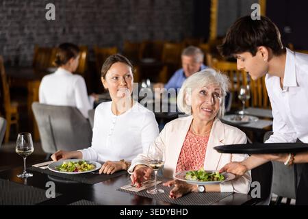 Jeune serveur servant des plats aux invités féminins dans le restaurant Banque D'Images