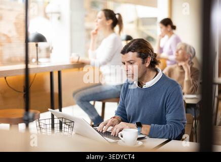 Homme concentré buvant du café tout en travaillant sur ordinateur portable dans un café confortable Banque D'Images
