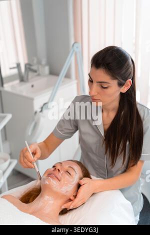 Heureux massothérapeute appliquant le masque facial à la femme avec brosse au salon de cosmétologie. La cliente féminine détendue se fait dorloter dans le spa par un professionnel Banque D'Images