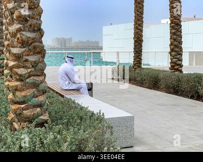 Vue d'un homme en vêtements traditionnels émiratis assis sur un banc le long d'une promenade au bord de l'eau avec des palmiers et des bâtiments modernes à Dubaï, Dubaï, ONU Banque D'Images