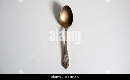 Une cuillère en métal repose à plat sur une surface blanche. Il a un look usé avec quelques détails sur la poignée. La cuillère est centrée et se détache contre la plaine Banque D'Images