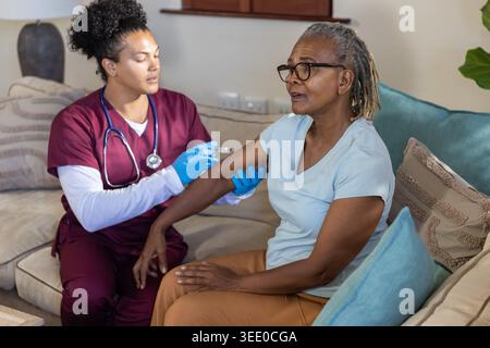 Femme afro-américaine senior vaccinée à domicile par une infirmière en gommage Bordeaux, gants bleus Banque D'Images