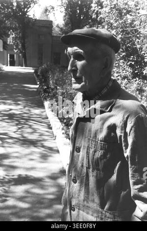 Sloviansk, 1997, Centre-ville. Portrait archivistique en noir et blanc d'un homme âgé debout à l'extérieur dans un parc ensoleillé. Il est habillé de façon décontractée dans une veste boutonnée sombre et une casquette plate traditionnelle, avec des traits du visage profondément doublés reflétant une longue expérience de vie. Le fond naturel présente une végétation luxuriante et des arbres en fleurs, ajoutant une atmosphère sereine à la composition. Cette étude de caractère franche capture l'esprit résilient et les visages quotidiens de l'ancienne génération dans le Donbass paisible. Banque D'Images