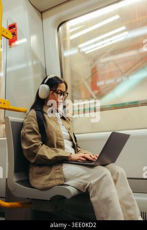 Un voyageur concentré utilise un ordinateur portable tout en portant des écouteurs dans un train de métro, tapant et travaillant à distance pendant son voyage et nous rendant productifs Banque D'Images