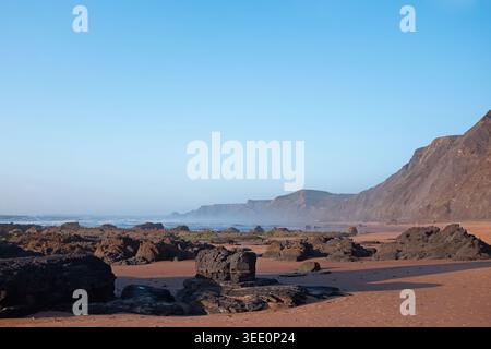 Algarve, Portugal : formations rocheuses sombres accidentées sur la plage de sable de Praia do Castelejo à marée basse. Espace de copie estival atmosphérique. Voyage en Europe contexte. Banque D'Images
