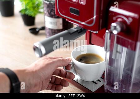 Gros plan d'une personne tenant une tasse blanche remplie de café expresso frais d'une machine à espresso rouge dans une cuisine moderne. Café maison confortable préparati Banque D'Images