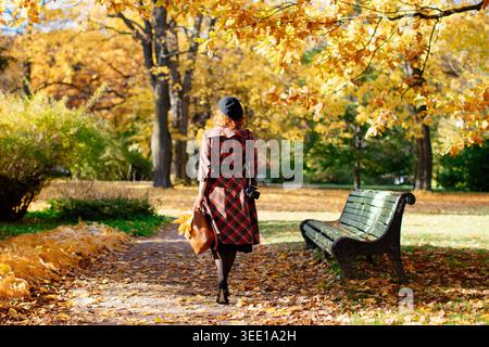 Élégante femme rousse en manteau à carreaux et béret noir promenades à travers le parc, tenant des feuilles d'érable sèches et sac à dos à la journée ensoleillée, vue arrière.humeur d'automne, concept de feuillage. Temps de détente dans le parc de la ville Banque D'Images