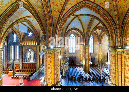 Les gens explorent l'église Matthias dans le château de Buda. L'intérieur coloré montre de hauts plafonds, des vitraux et des sièges en bois. Banque D'Images