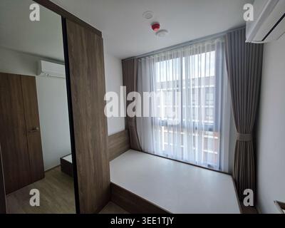 L'intérieur moderne de l'appartement de chambre vide dispose de meubles en bois près de la fenêtre lumineuse avec un rideau gris transparent offrant une atmosphère paisible et relaxante pour Banque D'Images