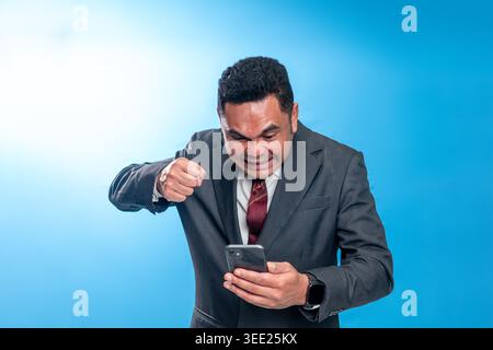 Portrait d'un cadre indonésien en colère en costume, serrant le poing et criant sur l'écran de son smartphone sur fond bleu. Banque D'Images