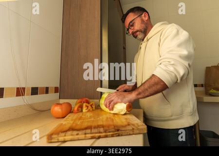 L'homme s'engage dans la préparation des aliments, épluchant méticuleusement et coupant les légumes frais sur une planche à découper en bois. Il met l'accent sur la promotion de la santé Banque D'Images