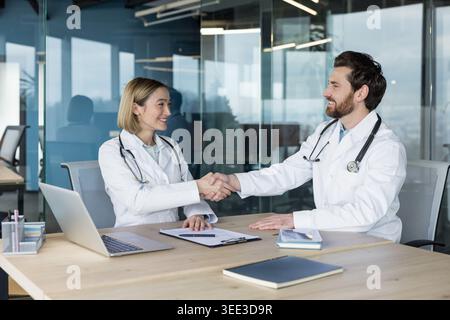 Deux médecins femme asiatique et homme caucasien en manteaux blancs avec stéthoscopes se serrent la main sur un bureau dans un bureau moderne, célébrant un partenariat médical réussi et la confiance Banque D'Images