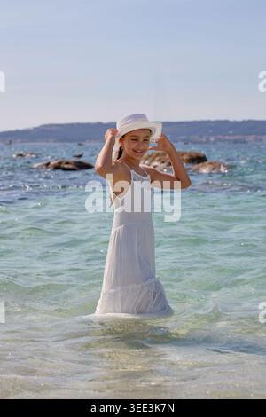 Femme debout dans l'eau de mer peu profonde à la plage tout en ajustant son chapeau. Banque D'Images