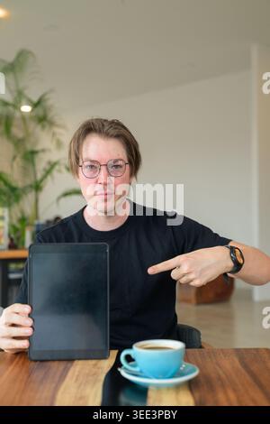 Homme scandinave assis à une table en bois dans un café lumineux, faisant des gestes et pointant son doigt vers un écran de tablette numérique vierge. C'est un adulte St Banque D'Images