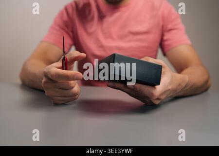 Mains de la personne utilisant le petit multi-outil rouge pour ouvrir, déballer et déballer le nouveau gadget de produit de la boîte d'emballage en carton noir assis à la table, révélant R Banque D'Images