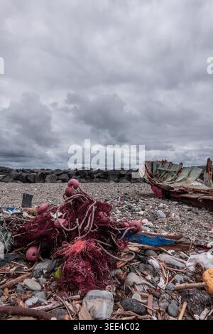Une vue rapprochée des filets de pêche rouges et des cordes avec bouées jetées, le long d'une épave de bateau délabrée, reposant sur une plage rocheuse et parsemée de débris. Th Banque D'Images