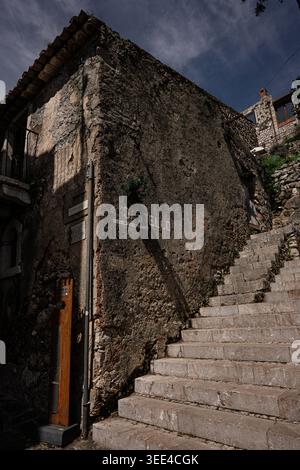 Un bâtiment en pierre vieillie avec un toit rustique en tuiles, doté d'un balcon étroit, se dresse à côté d'un escalier en pierre menant vers le haut. Le bâtiment est texturé Banque D'Images