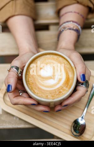 Les mains avec le vernis à ongles bleu et les anneaux cachent une tasse à latte chaude surmontée d'art en forme de coeur sur un plateau en bois dans un cadre de CAF confortable, évoquant comfo Banque D'Images