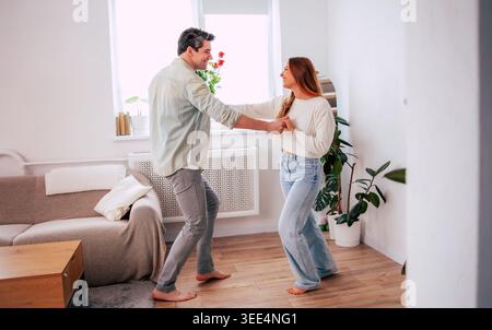 Heureux jeune couple dansant ensemble à la maison, homme romantique tenant des roses rouges et femme surprenante dans un salon moderne lumineux Banque D'Images