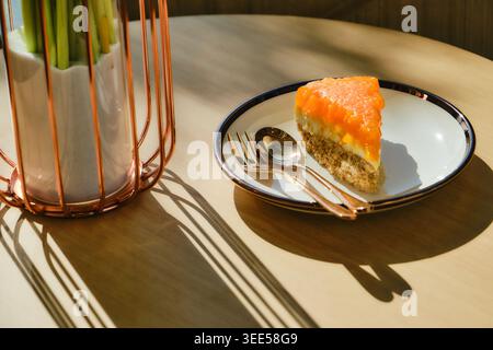 Une tranche de gâteau à l'orange, assise sur un plat, est servie sur une table dans un café à côté d'un vase de fleurs. La douce lumière du soleil de l'après-midi pénètre à travers le th Banque D'Images
