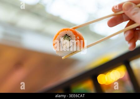 Gros plan d'un rouleau de saumon frais tenu avec des baguettes dans un restaurant accueillant. Le poisson orange éclatant recouvre un riz parfaitement formé avec un style propre et appétissant. Belle vitrine de sushi à son meilleur Banque D'Images