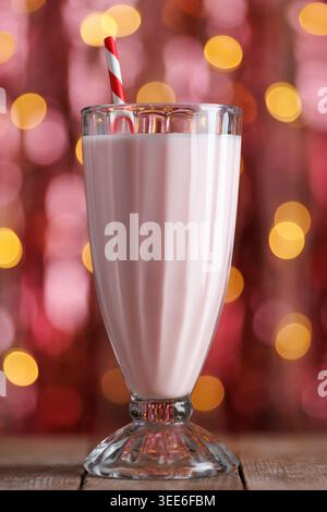 Milk-shake aux fraises avec paille rouge rayée dans un grand verre assis sur une table en bois avec des lumières bokeh étincelantes Banque D'Images
