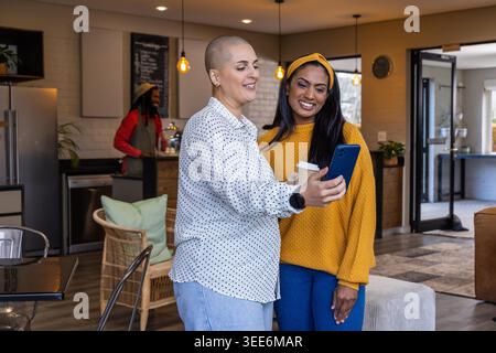 Diverses amies féminines debout, vérifiant le téléphone bleu dans le salon avec du café, portant un pull moutarde. Café, coworking, chaleur, moderne, collaboration, connexion, plantes Banque D'Images
