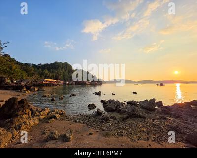 Coucher de soleil paisible sur la plage de l'île tropicale avec rivage rocheux eau de mer calme reflet du soleil ciel bleu clair petits bateaux distance créant Ser Banque D'Images
