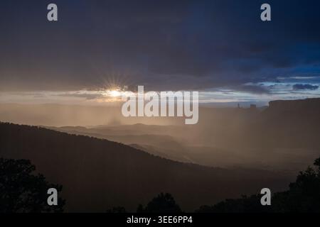 Vue d'un soleil éclatant à travers les nuages sombres de tempête au-dessus d'une vallée désertique brumeuse avec des collines silhouettées à Moab, Utah, États-Unis. Banque D'Images