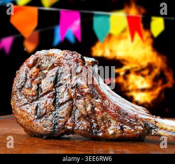 Steak tomahawk grillé sur une planche de bois avec des décorations traditionnelles brésiliennes Festa Junina, des drapeaux colorés et une flamme de feu de joie dans le dos flou Banque D'Images
