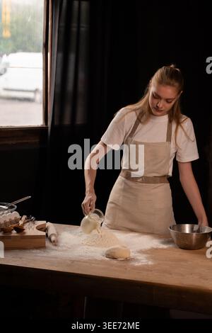 Femme versant du lait dans la farine sur une table en bois, préparant la pâte pour faire cuire le pain, cuisiner dans la cuisine Banque D'Images