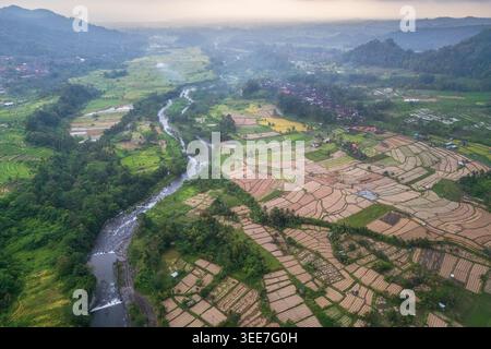 Vue aérienne de rizières luxuriantes en terrasses et d'une rivière sinueuse qui coule à travers une vallée tropicale sous un ciel brumeux à Sidemen, Bali, Indonésie. Banque D'Images