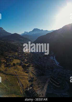 Vue aérienne du village alpin de Pie' Falcade niché dans une vallée entourée par les majestueux sommets enneigés des Dolomites sous un soleil éclatant Banque D'Images