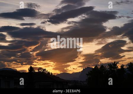Un coucher de soleil orange spectaculaire illumine des nuages lenticulaires au-dessus des toits, des arbres et des sommets montagneux lointains. Banque D'Images