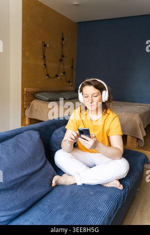 Femme assise les jambes croisées sur un canapé confortable, portant des écouteurs et utilisant son smartphone pour se divertir, surfant sur Internet sur son téléphone Banque D'Images