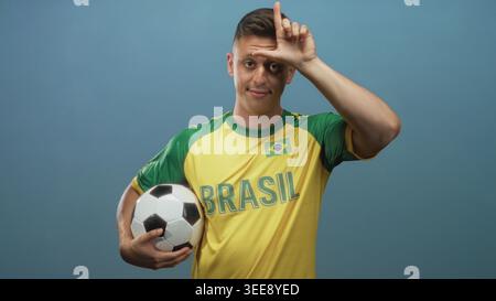 Jeune homme tient le ballon de football et fait l signe sur le front en studio tout en portant le maillot jaune du brésil avec patch drapeau et cheveux blonds ; rivalité moqueuse. Banque D'Images