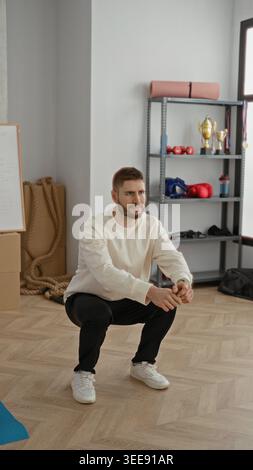 Homme squattant avec les mains serrées et des baskets blanches sur plancher de bois dans un petit studio de fitness avec étagère de trophées, haltères, gants de boxe et battl Banque D'Images