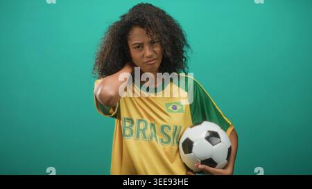 Adolescente tenant le ballon de football tient le cou en studio, portant le t-shirt de l'équipe du brésil et griffant avec les yeux fermés ; résilience de l'inconfort. Banque D'Images