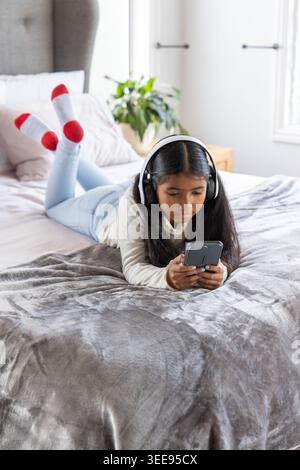 Fille indienne allongée sur le lit dans une chambre lumineuse portant des écouteurs et tenant un smartphone avec support Banque D'Images