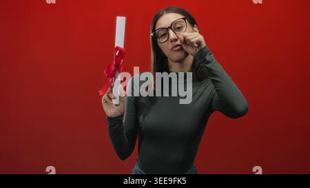Jeune femme portant des lunettes tient un rouleau de diplôme noué avec un ruban rouge et se frotte les yeux tout en posant sur un fond de studio rouge ; fierté su Banque D'Images