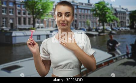 Femme aux cheveux courts tient un rasoir rose dans sa main droite tout en touchant la poitrine avec la main gauche sur une rue du canal par des bateaux et des bâtiments en briques ; hesitat Banque D'Images