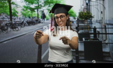 Femme portant une casquette de remise des diplômes pointe les doigts avec les deux mains vers la caméra dans la rue près des vélos et des rampes, souriant et se faisant passer pour un étudiant ; achie Banque D'Images