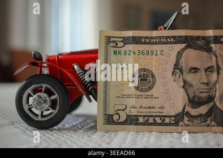 Une voiture rouge vintage exposée à côté d'un billet de cinq dollars des États-Unis sur une surface texturée blanche. Cette image conceptuelle représente le contraste Banque D'Images