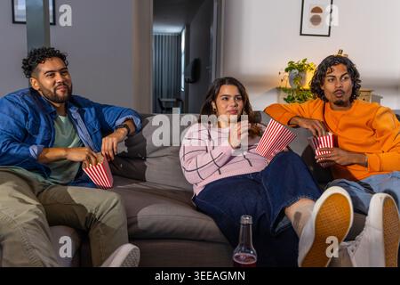 Divers amis assis sur le canapé regardant un film dans le salon tenant des boîtes de pop-corn rayées Banque D'Images