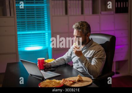 Freelance manger de la pizza tard le soir. Bureau de nuit avec ordinateur portable et collation. Détente au bureau de nuit. Travail de nuit. Homme d'affaires mangent collation tout en travaillant sur Banque D'Images