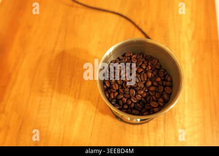 Grains de café remplissant un moulin électrique, préparant du café frais. Prise de vue aérienne, lumière chaude du matin sur une surface en bois Banque D'Images