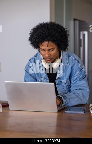 Femme afro-américaine tapant sur un ordinateur portable argenté à la table de cuisine portant des écouteurs de veste en denim. Espace de travail, télétravail, bureau à domicile, tablette, téléphone, casque, minimaliste Banque D'Images