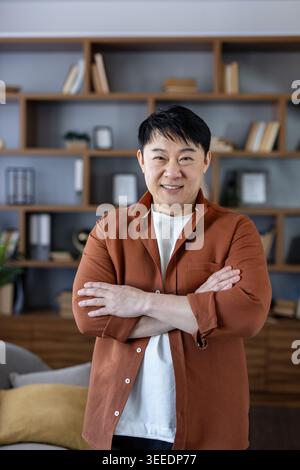 Homme asiatique d'âge moyen confiant souriant, debout avec les bras croisés dans un bureau à domicile ou un salon moderne, représentant des concepts de réussite, d'éducation et de travail à distance heureux Banque D'Images