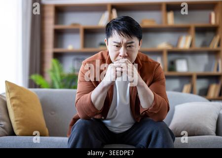 Homme asiatique se concentrant profondément sur un problème, éprouvant du stress ou de l'anxiété tout en étant assis sur un canapé dans son salon moderne, réfléchissant sur la solitude ou la santé mentale Banque D'Images