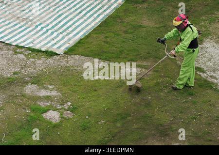 Plan surélevé d'un technicien paysagiste utilisant une tondeuse sur de l'herbe patchy, portant une combinaison et un chapeau fluo, stable Banque D'Images